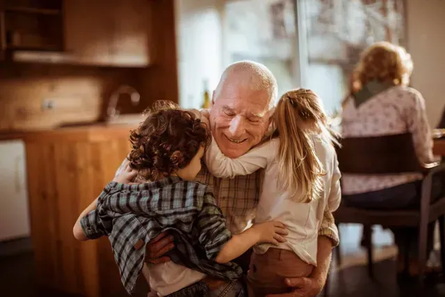 A smiling elderly person hugs two children in a warm kitchen while another person sits at a table nearby.