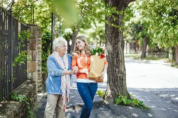 Two people walking on a tree-lined sidewalk, carrying grocery bags and smiling together