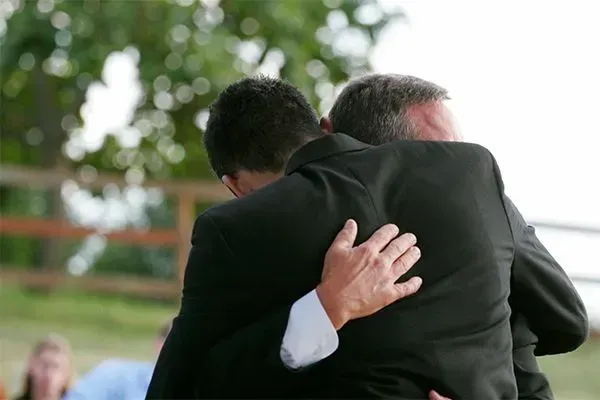 Two people in dark suits embrace in a close hug outdoors, with trees blurred in the background.