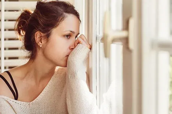 Woman in a beige sweater leaning by a window, looking contemplative with her hand near her face