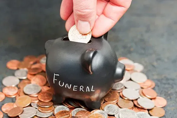 Hand placing a coin into a black piggy bank labeled “Funeral,” surrounded by scattered coins.