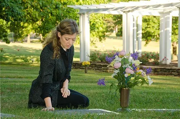 Person kneeling by a grave in a green cemetery beside a bouquet of purple flowers