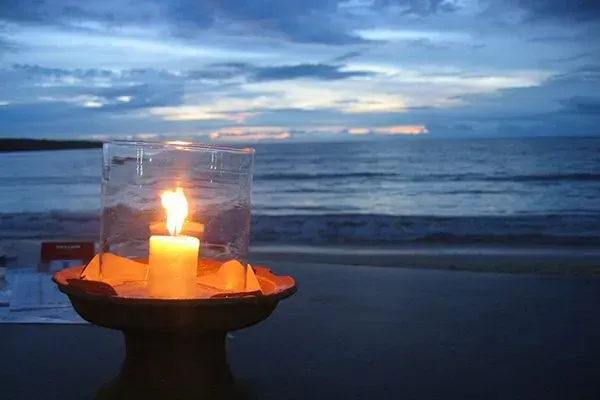 Lit candle in a glass holder on a beach at dusk, with waves and cloudy sky in the background