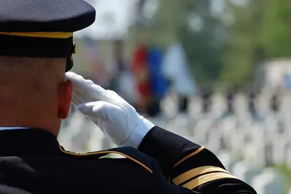 Uniformed soldier saluting at a memorial with rows of white headstones in the background