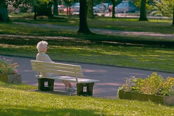 Person sitting on a park bench beside a paved path, surrounded by green grass and trees.