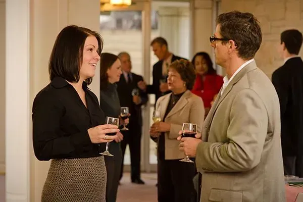 Two people chatting with drinks at a formal indoor reception, with guests mingling in the background
