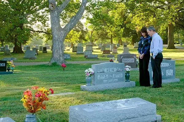 Two people stand beside a gravestone in a cemetery with trees and flowers nearby.