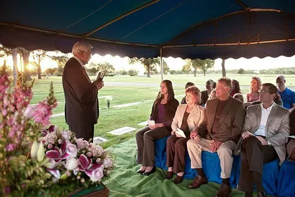 Outdoor ceremony under a tent, speaker addressing seated guests beside a flower arrangement.