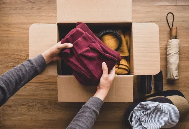 Hands packing a burgundy sweater into a cardboard box with yarn and folded clothes on a wooden table