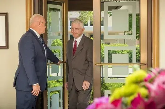 Two men shake hands at a doorway, with flowers blurred in the foreground.