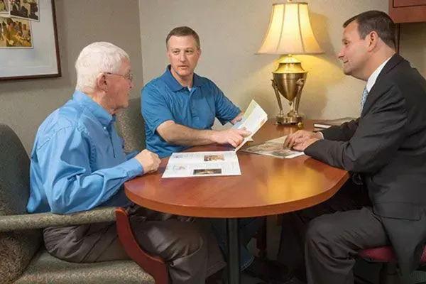 Three men sit at a round table in a meeting room, reviewing papers beside a lamp.