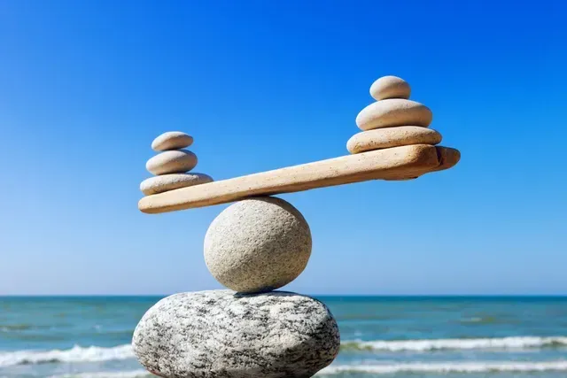 Balanced stones on a beach, with pebbles on a wooden plank against a blue sky.
