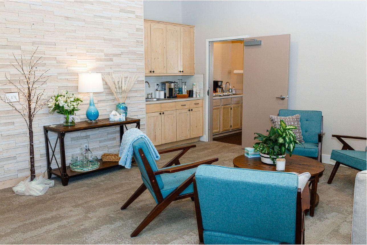 Bright living room with teal chairs, wooden table, cabinets, flowers, and a white brick fireplace.