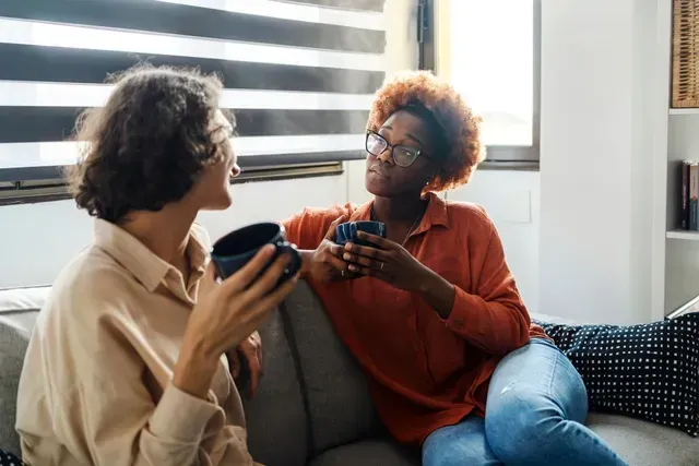 Two people on a couch holding game controllers and talking in a bright living room