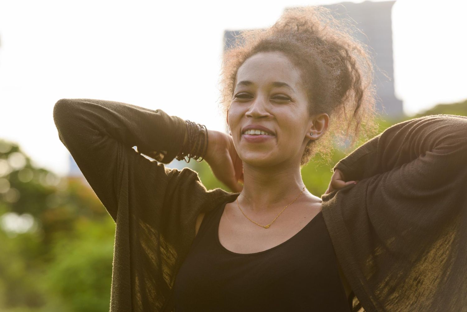 A woman with curly hair is standing in a park with her arms outstretched.