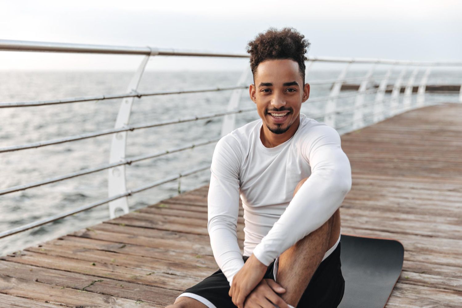 A man is sitting on a yoga mat on a pier near the ocean.