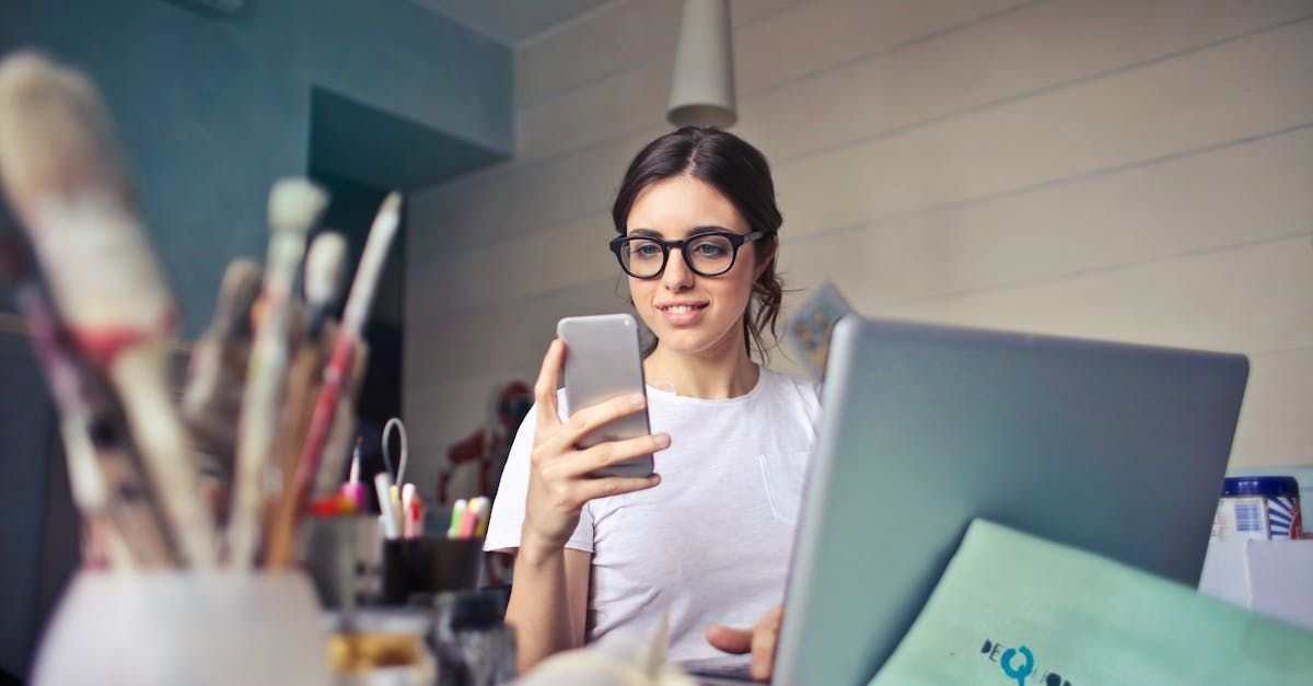 A woman is sitting at a desk using a laptop and a cell phone.