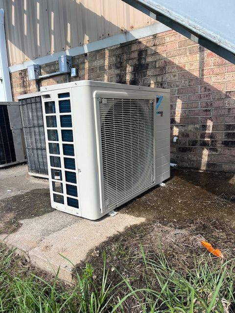 An air conditioning unit on a concrete pad, next to a brick building, with grass and dirt nearby.