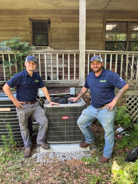 Two men in uniform stand with AC unit in front of a wooden house.