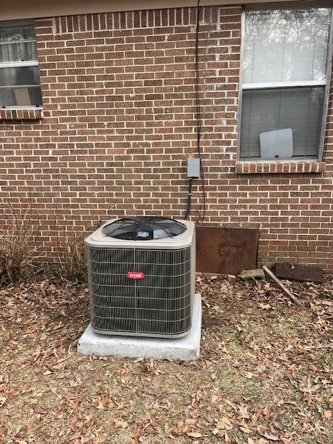 An air conditioning unit sits on a concrete pad outside a brick building next to a window.
