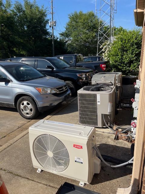 An air conditioning unit sits outside with other units near parked cars and a building on a sunny day.