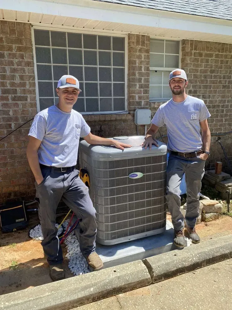 Two men are standing next to an air conditioner in front of a brick house.