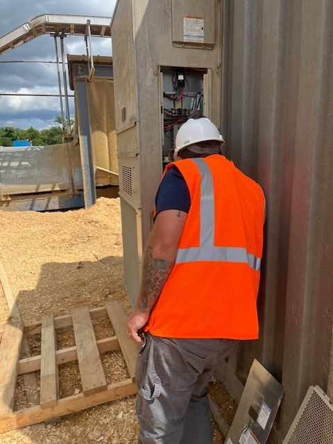 Construction worker in orange vest, helmet, leaning over electrical panel. Outdoors, gravel, conveyor belt in background.