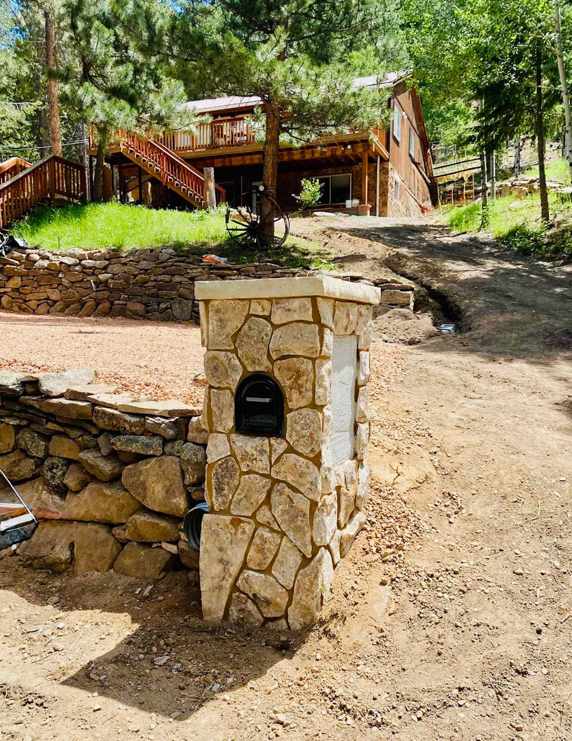 A stone mailbox is sitting in the dirt in front of a house.