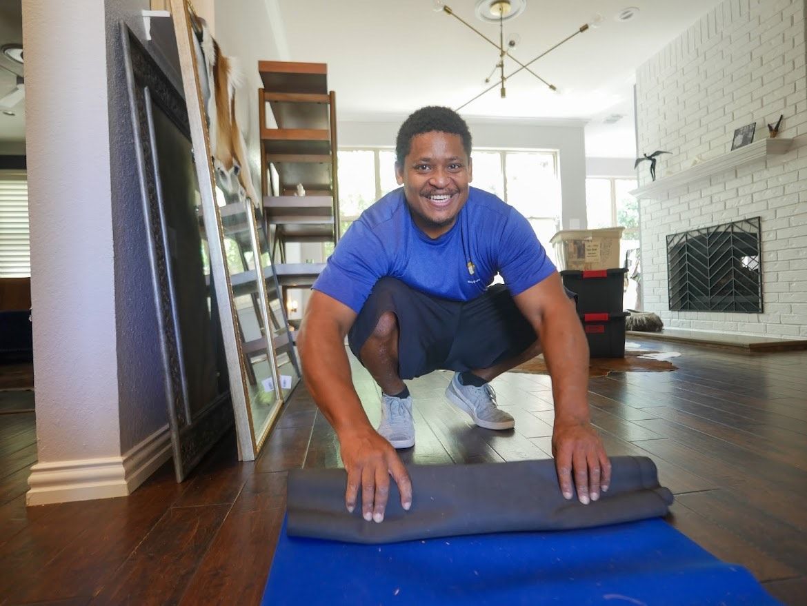 A man is squatting on a blue yoga mat in a living room.