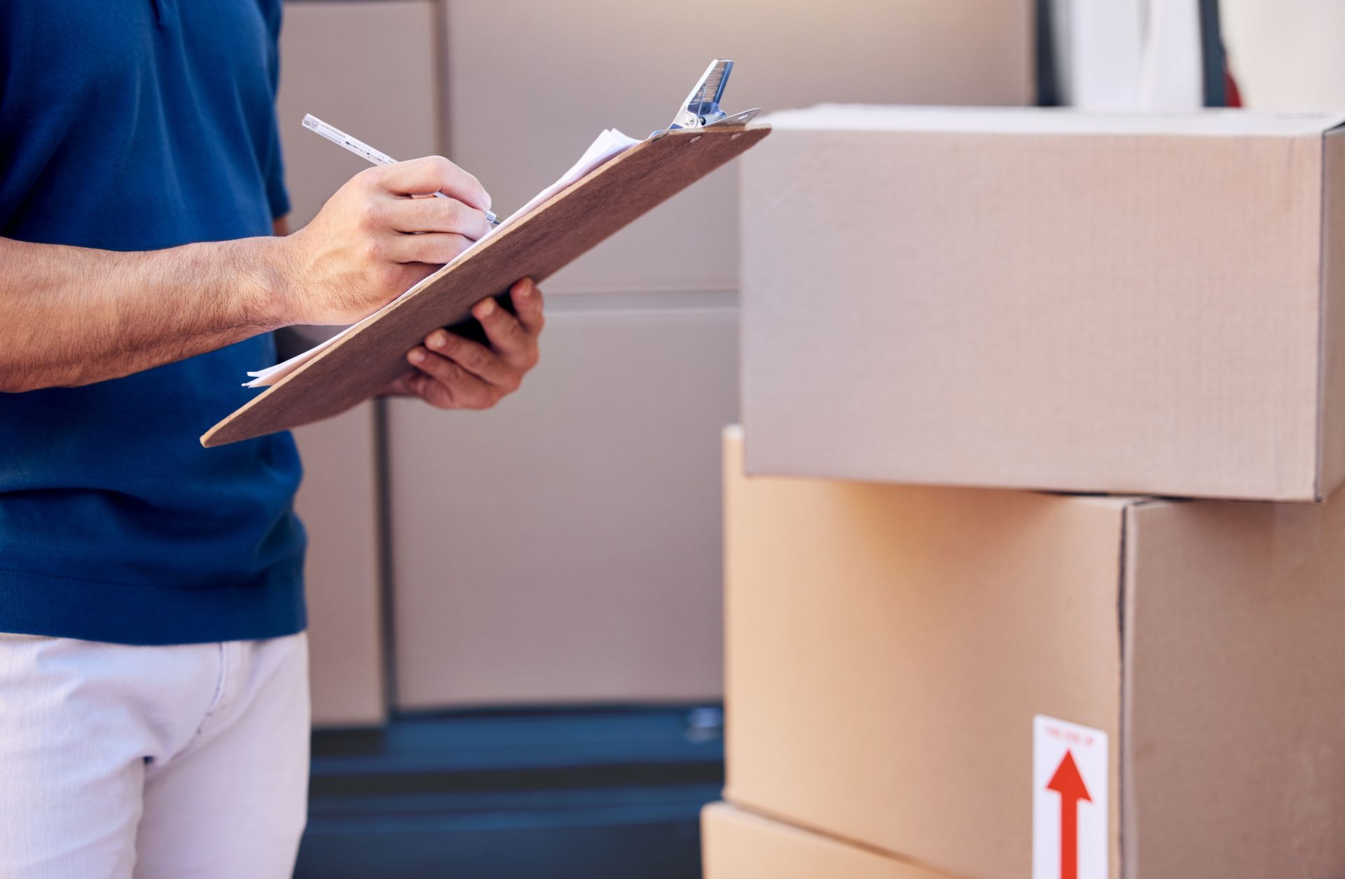 Person in blue shirt writing on clipboard next to stacked cardboard boxes in a truck.
