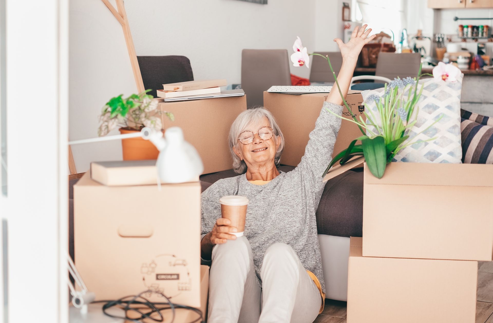 Elderly woman smiles and raises arm in excitement, sitting among moving boxes with coffee cup.