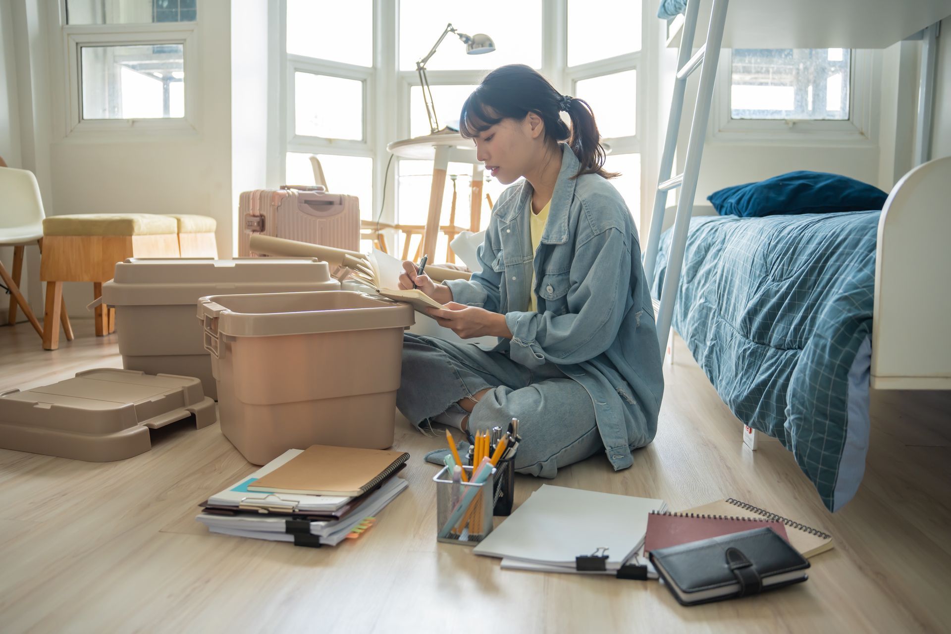Woman sitting on floor sorting through items, surrounded by boxes in a brightly lit bedroom.