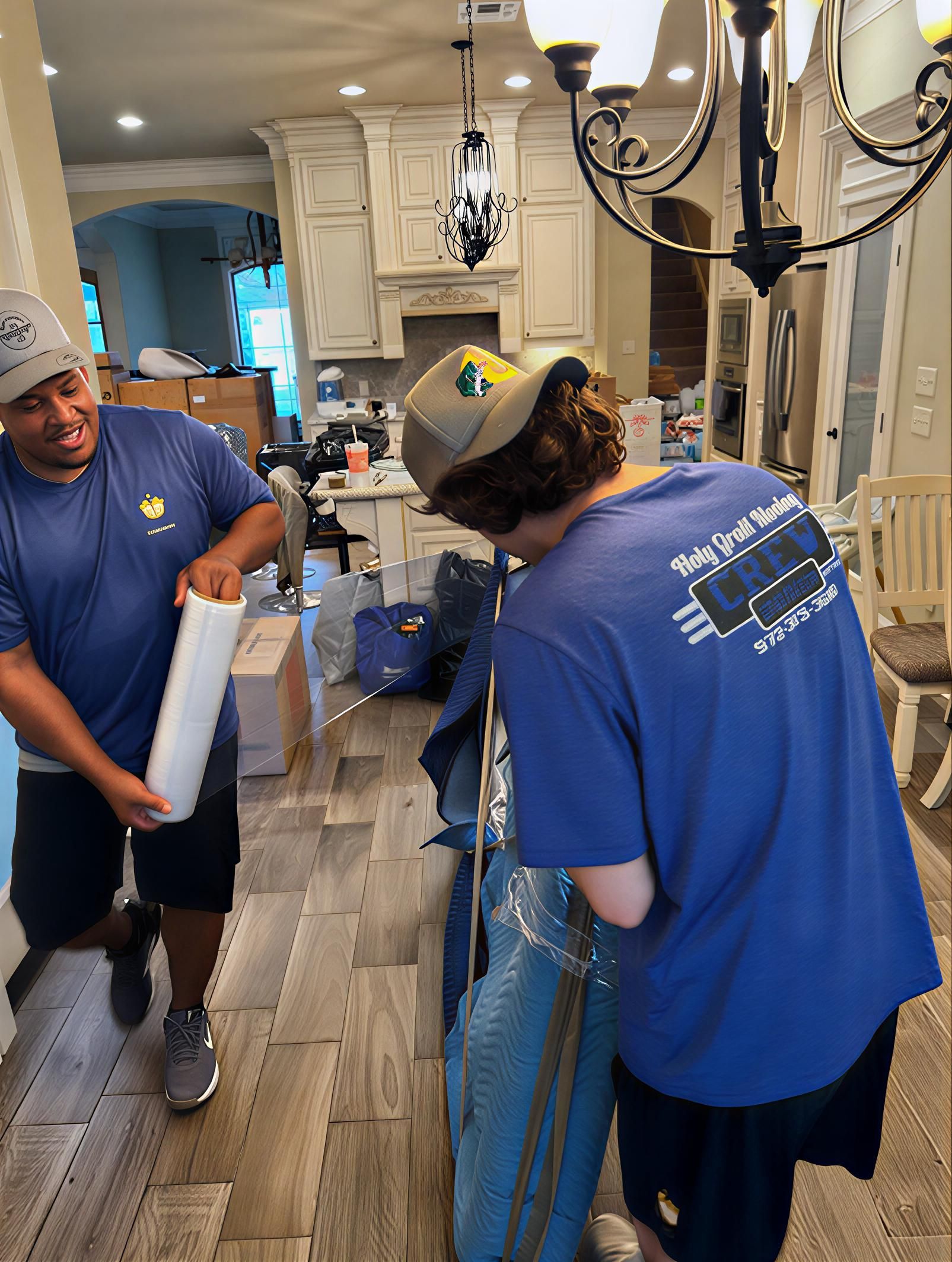 Two movers in blue shirts wrapping items inside a home with white cabinets and a chandelier.