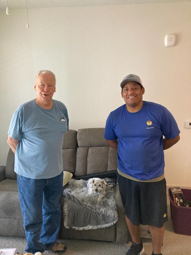 Two men are standing next to each other in a living room with a dog on the couch.