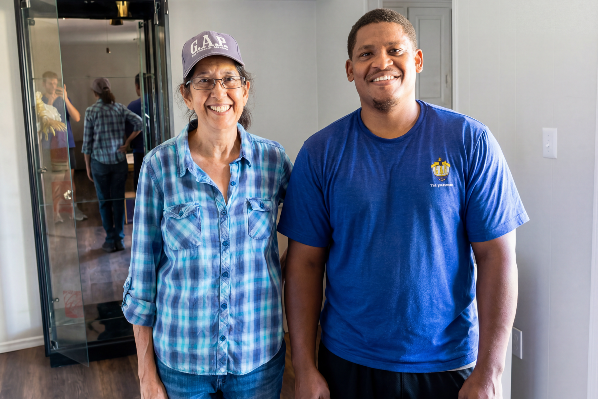 Woman and man standing side by side smiling. Large mirror behind them reflects people.