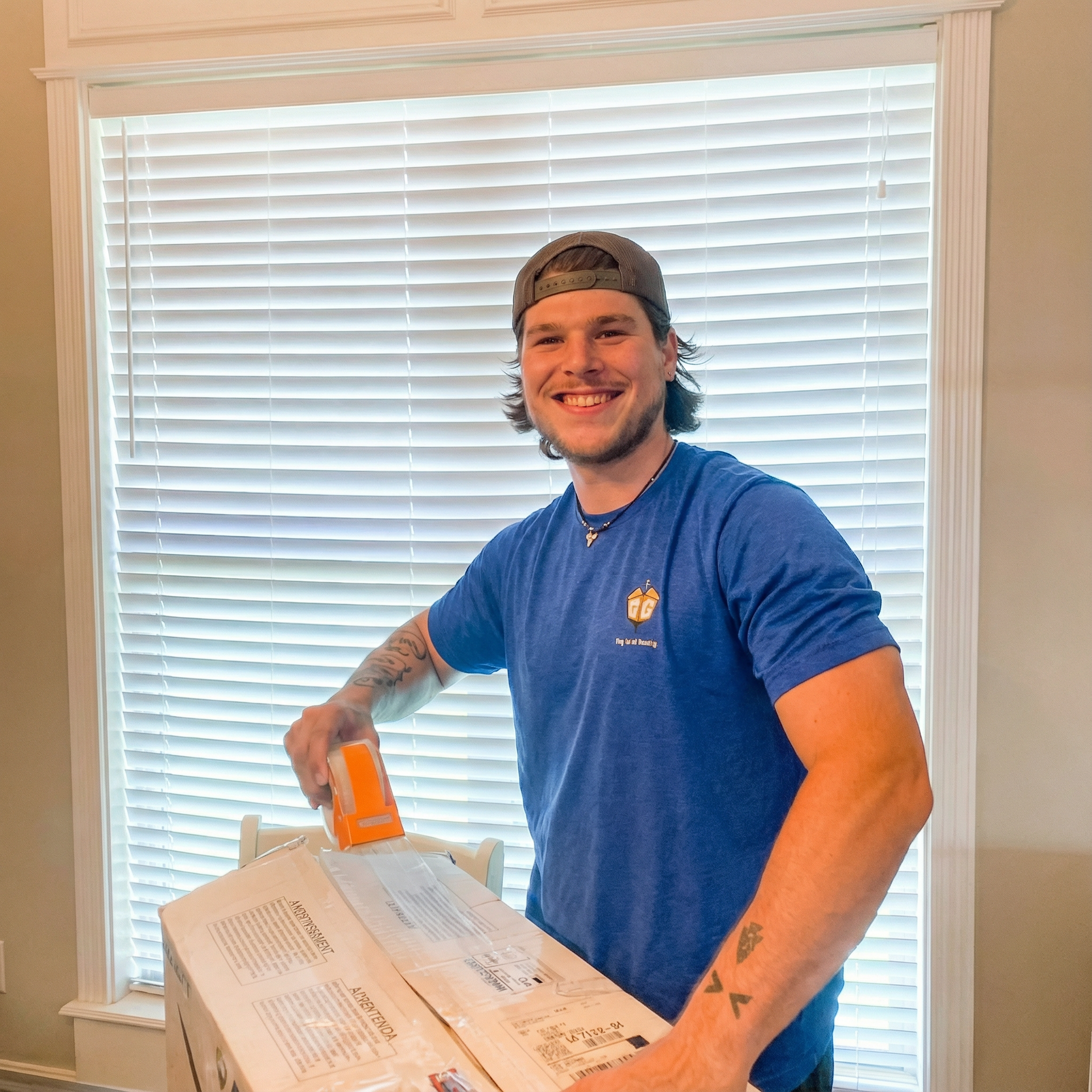 Man taping a cardboard box near a window, smiling. He wears a blue shirt and baseball cap.