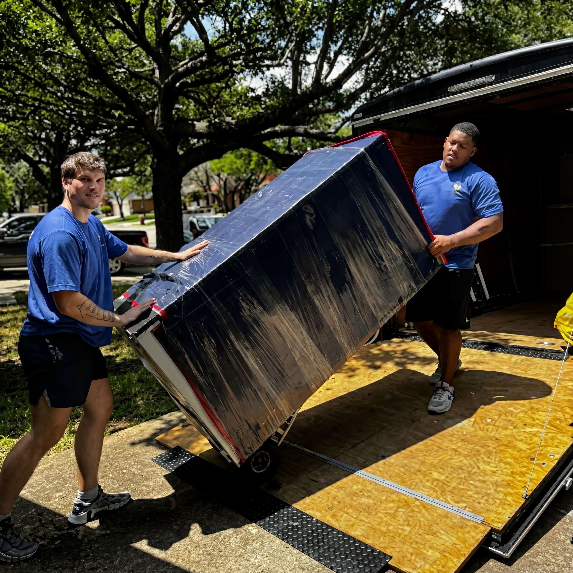 Two movers in blue shirts load a large box onto a truck; sunny outdoor setting.