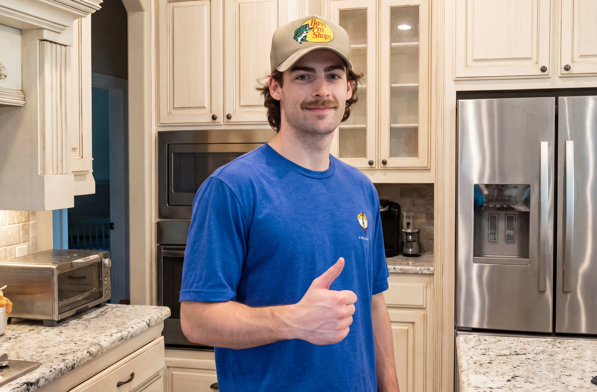 Man in blue shirt and Bass Pro hat gives a thumbs up in a kitchen.