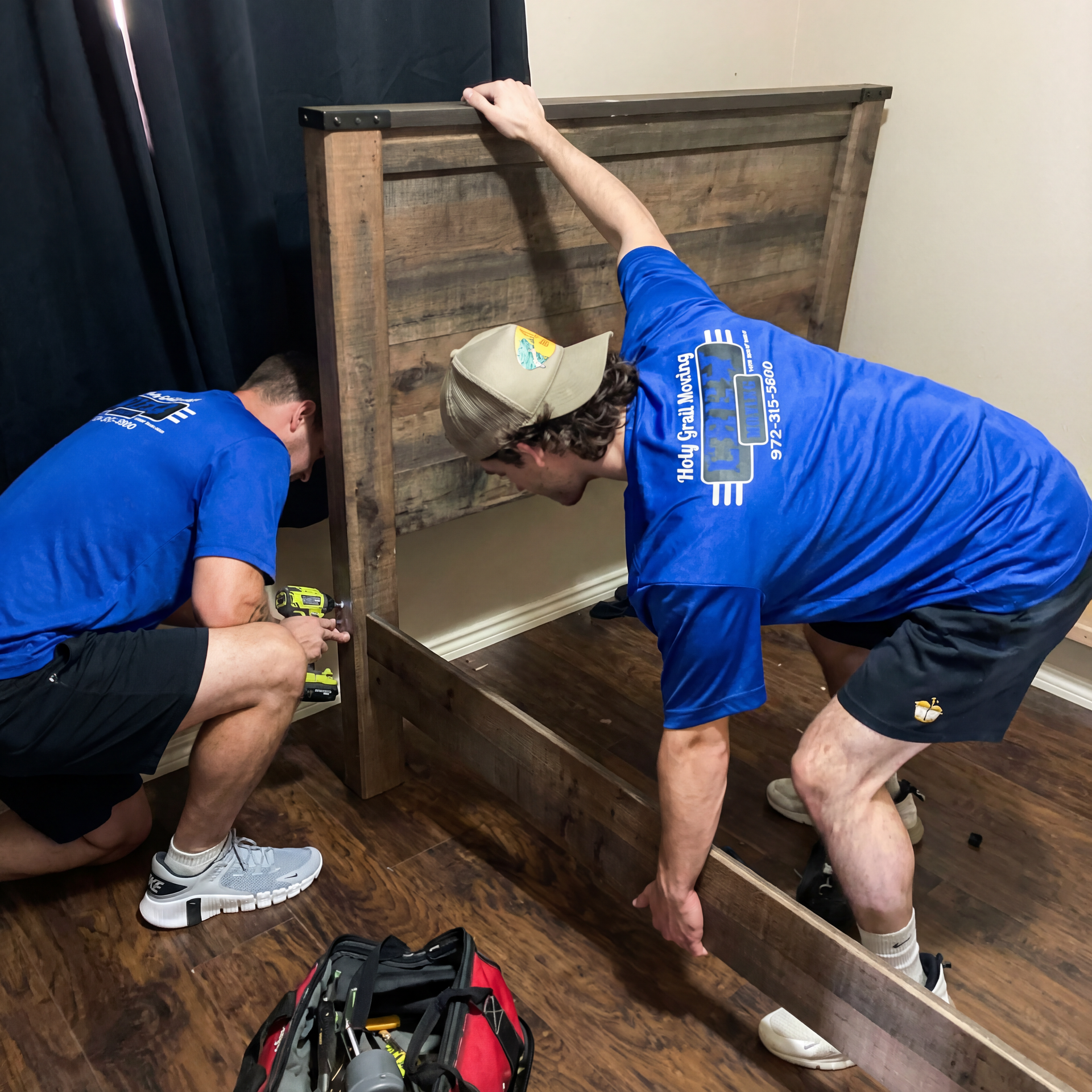 Two people in blue shirts assembling a wooden bed frame in a room. One uses a drill.
