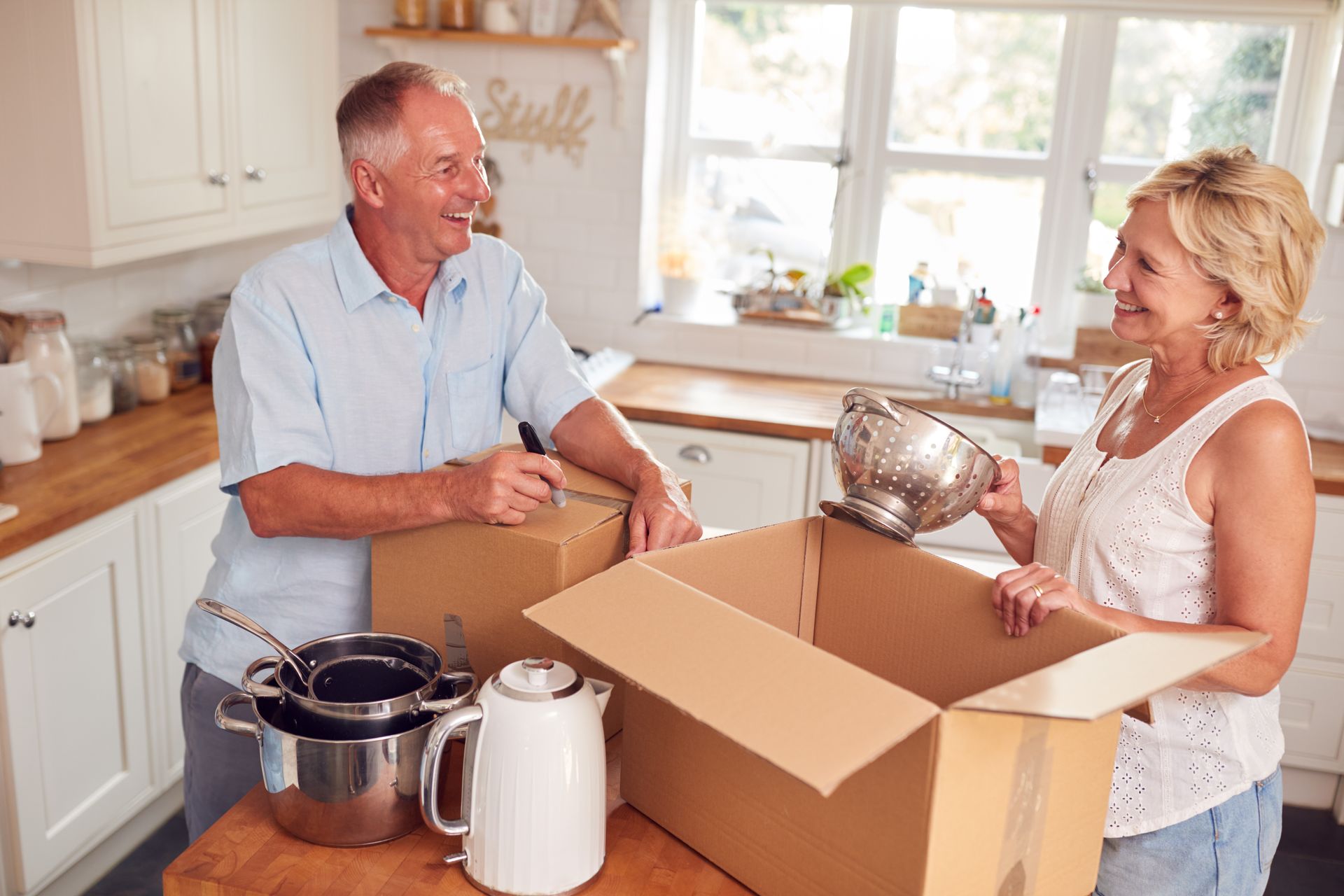 Couple packing kitchen items into cardboard boxes in a bright kitchen.