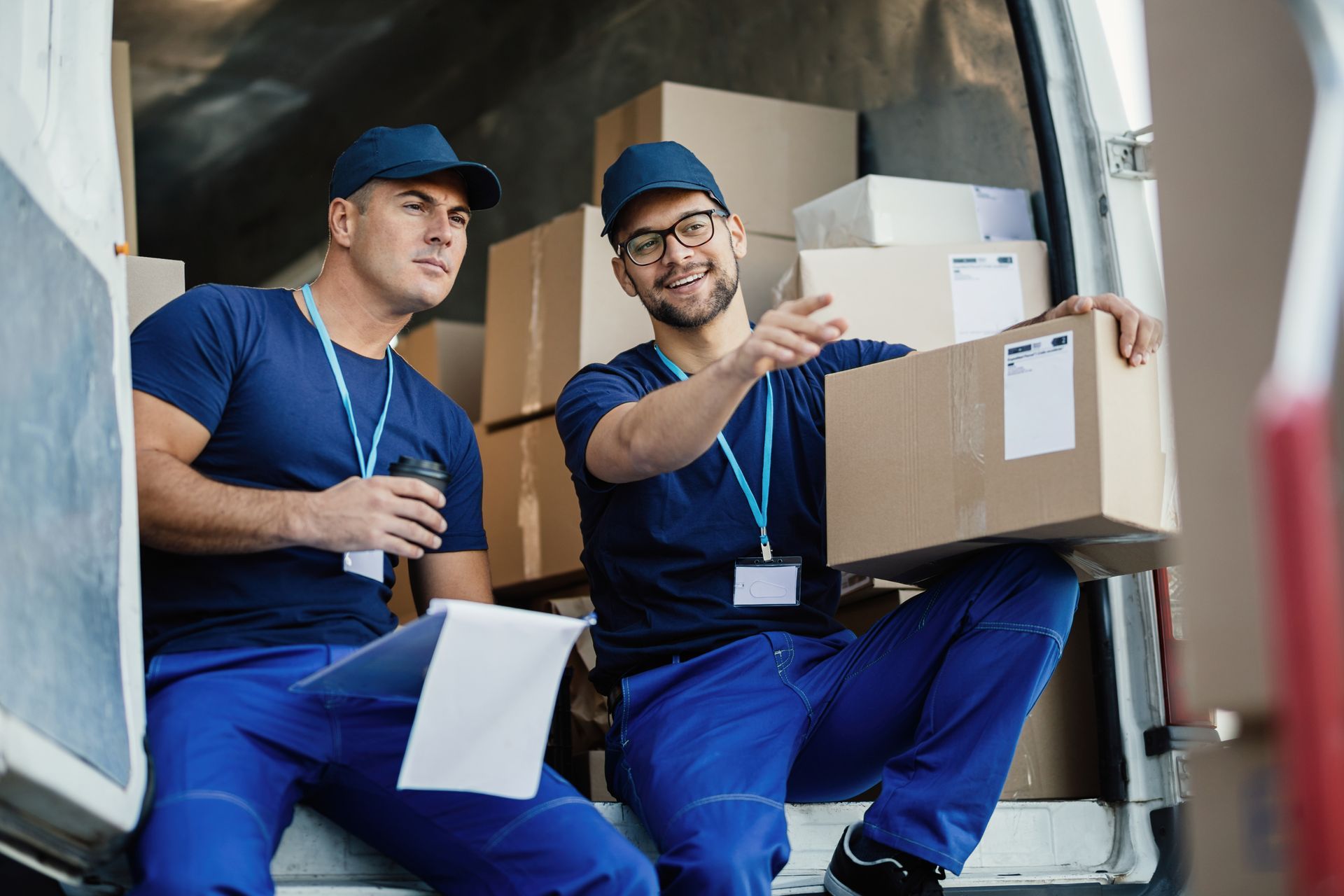 Two delivery workers in blue uniforms inside a van, one holding a box and pointing, the other holding papers.