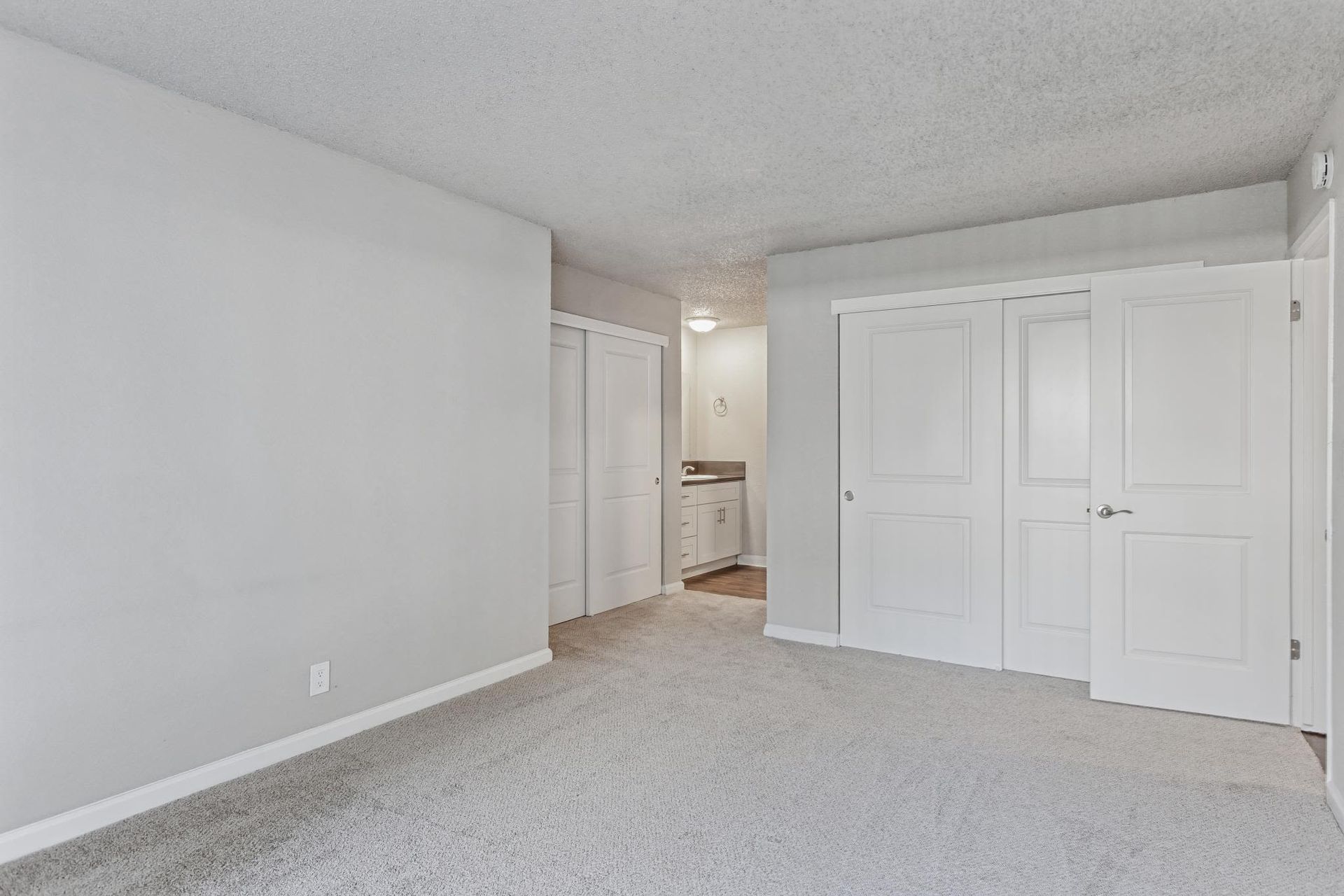 Bedroom with gray walls, white double closet doors, and beige carpet; view toward a small vanity area.