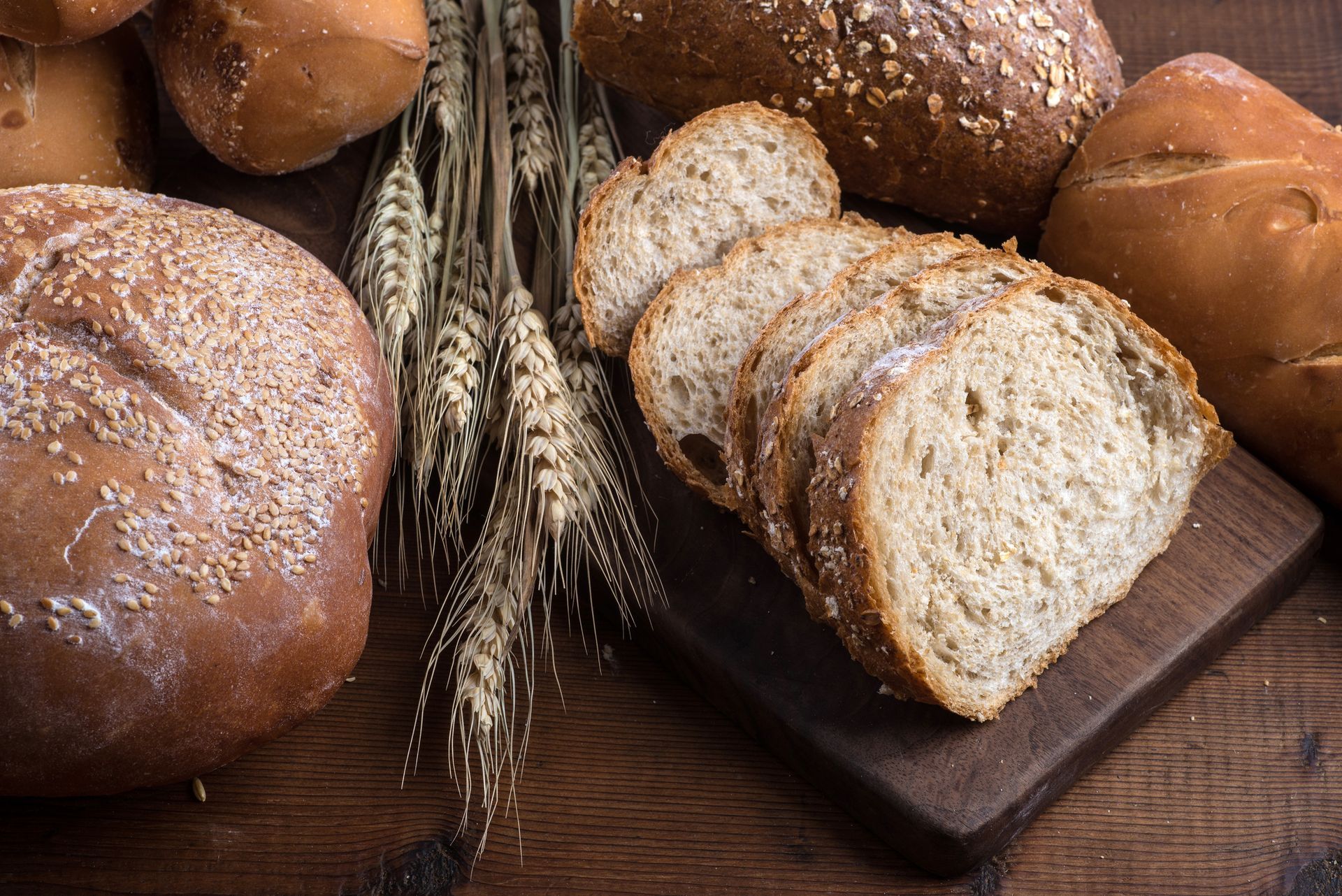 Sliced whole wheat bread on a wooden board, surrounded by loaves and wheat stalks.