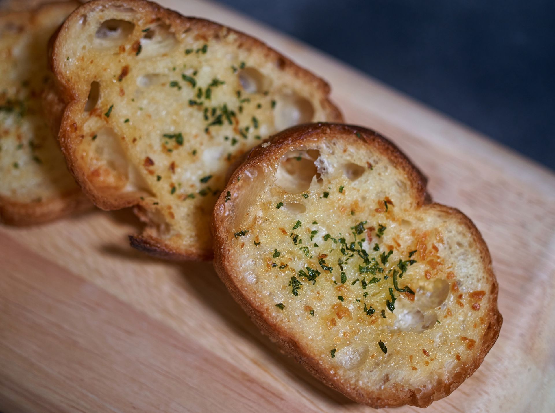 Garlic bread slices on a wooden board, golden brown with herbs.