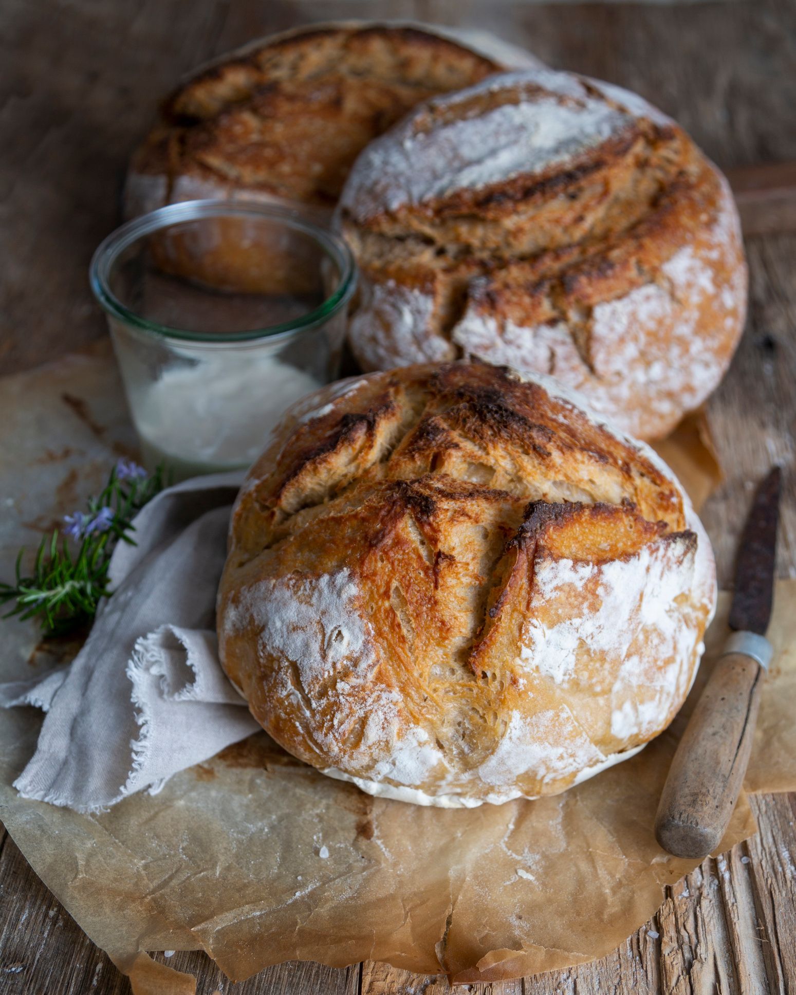 Three crusty loaves of bread on parchment paper with a jar of white spread and knife.