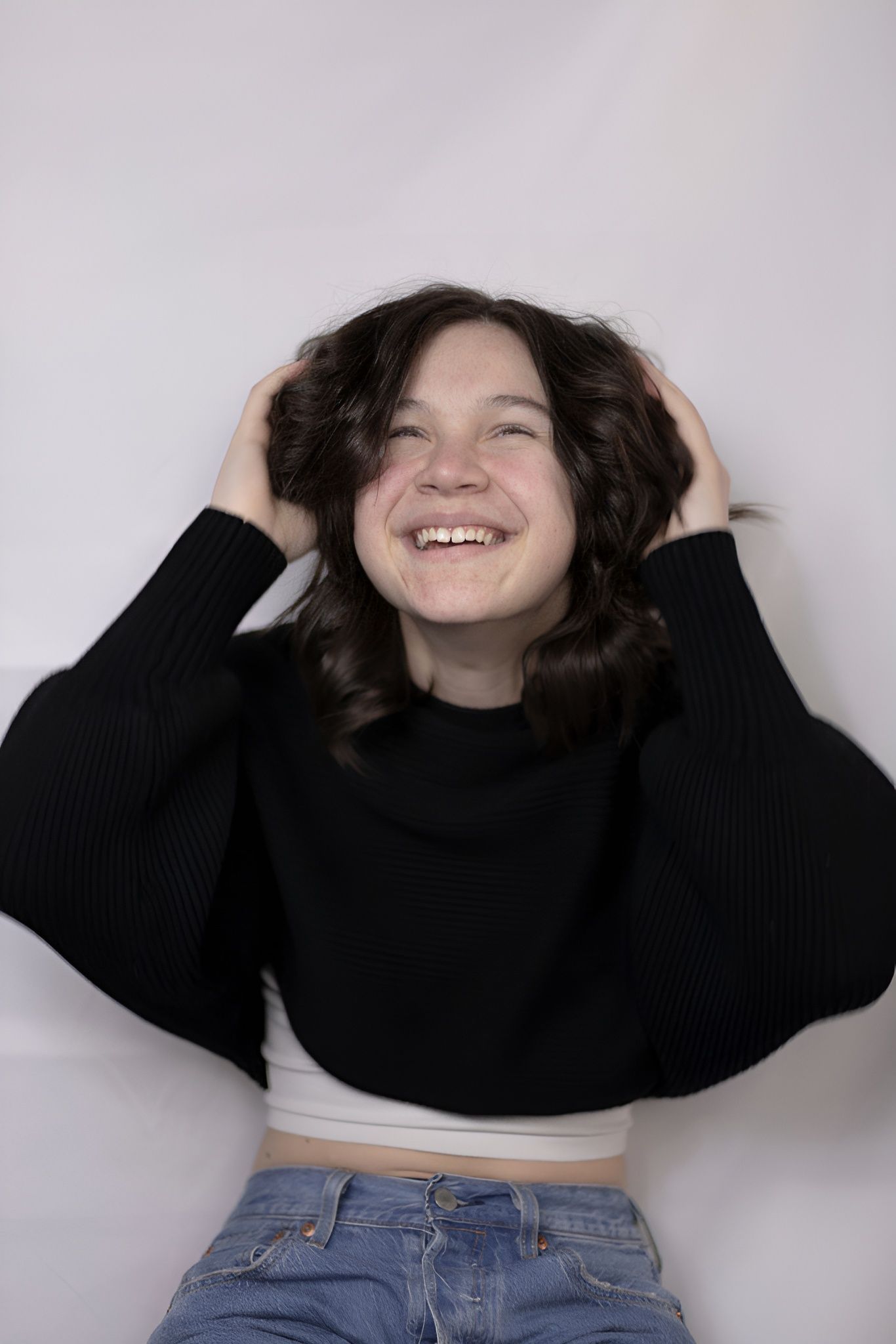 A young woman in a black sweater and blue jeans is smiling and holding her hair.