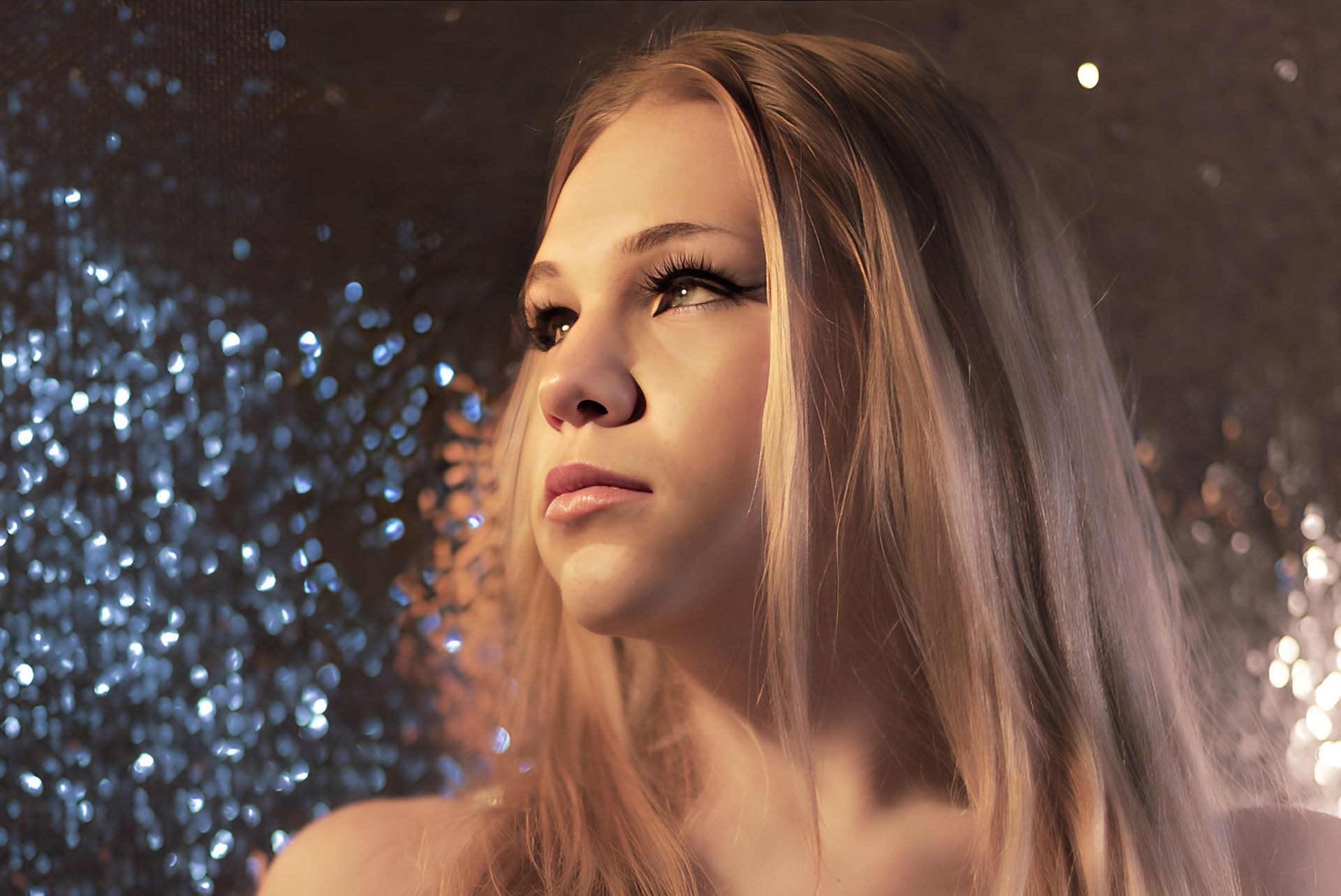A woman with long blonde hair is standing in front of a glittery background.