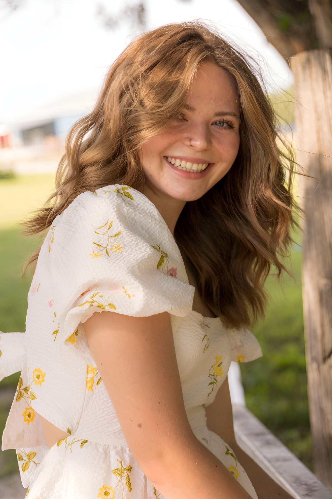 A woman in a white dress with yellow flowers is smiling while sitting under a tree.