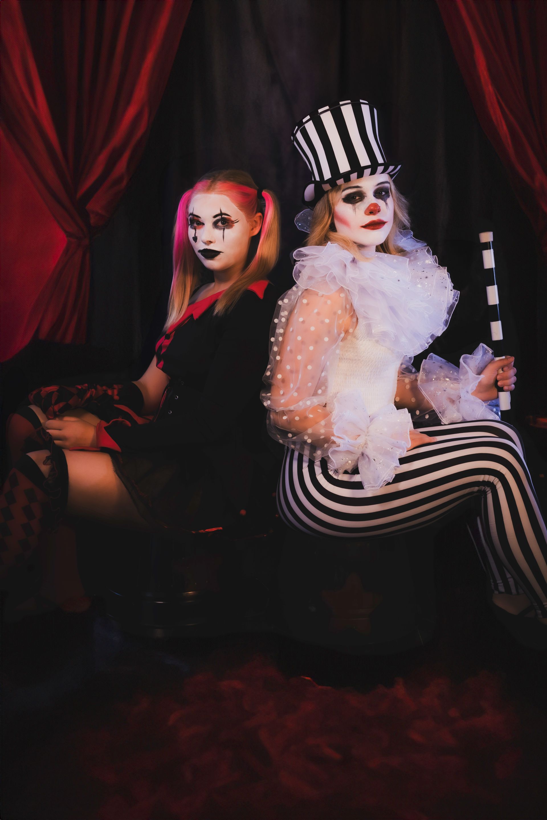 Two women dressed as clowns are sitting back to back in a room.