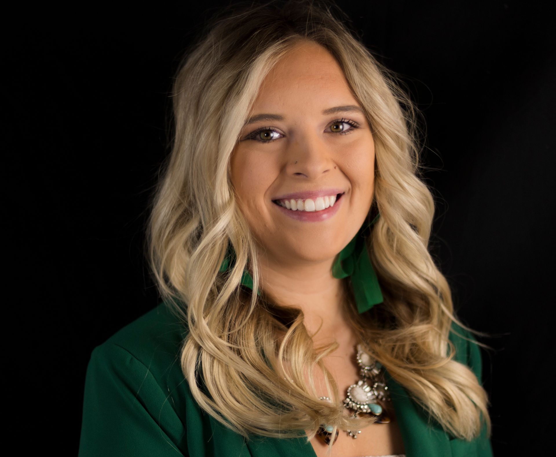 A woman wearing a green jacket and a necklace is smiling for the camera.